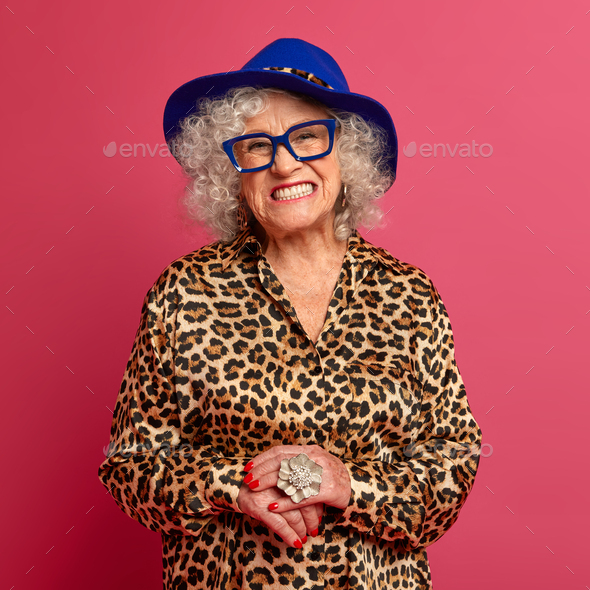 Happy grandmother smiles toothily, wears fashionable blue hat and leopard outfit, looks through tran