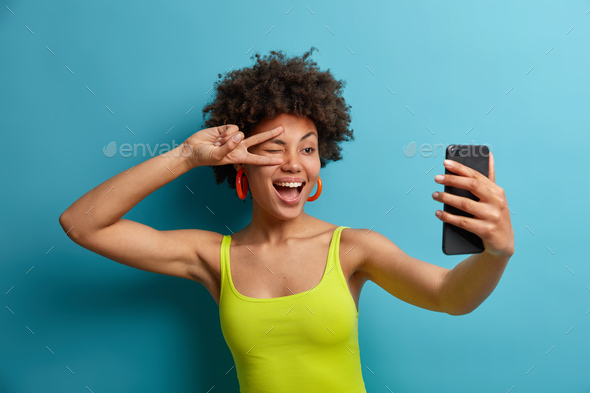 Playful positive curly haired woman makes peace sign over eye, takes selfie on smartphone, shows luc