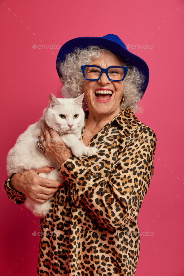 Elderly woman has happy mood, dressed elegantly, holds white cat on hands, ready for outdoor walk, f