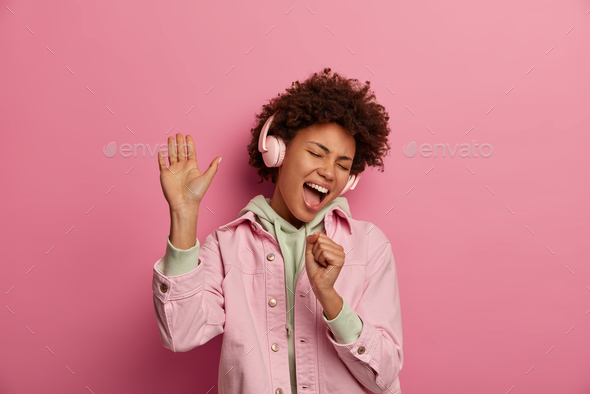 Photo of overjoyed woman sings along in headphones, makes microphone with hand, raises palm and danc