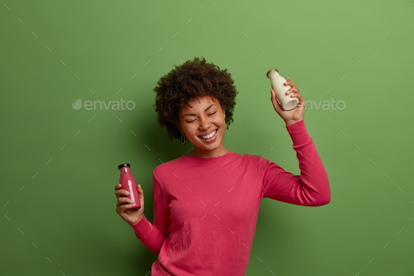Isolated shot of happy pleased Afro American woman poses with vegetarian milk and smoothie, enriches