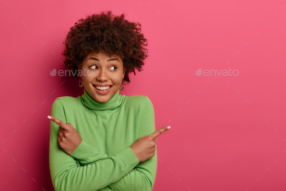 Cheerful curly haired woman crosses arms over chest, points left and ...