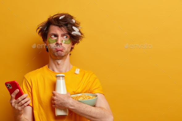 Morning and bad mood. Displeased man poses with ingredients for breakfast, scrolls news via smartpho