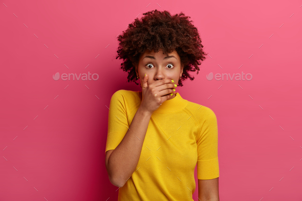 Scared dark skinned woman covers mouth, holds breath from fright, stands speechless, finds out shock