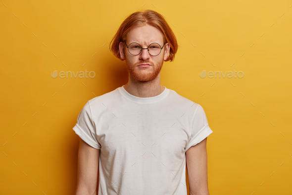 Horizontal shot of dissatisfied bearded ginger man smirks face, feels depressed and worried, frowns