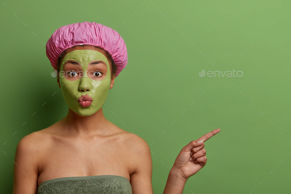 Lovely ethnic woman with folded lips, applies cosmetic mask on face, removes bleakheads, wears bath