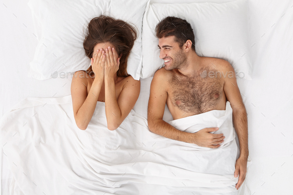 Happy lovers stay in cozy bed, have good mood after passionate sex, shy woman hides face with hands,