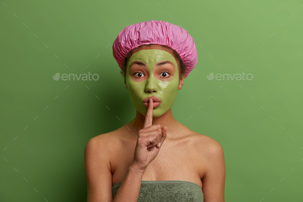 Surprised young female model with green peel off mask on her face, makes silence gesture, tells secr