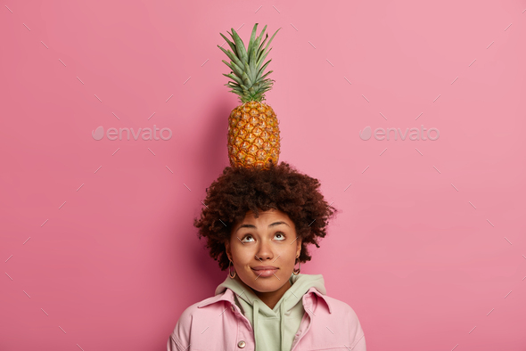 Pretty curly woman looks above, keeps balance with pineapple on head, concentrated above, has dark s