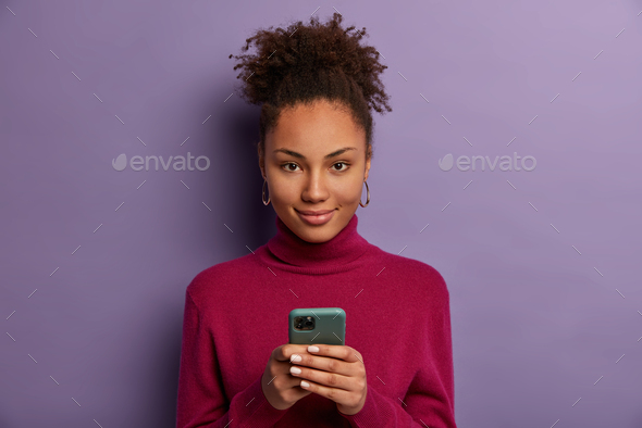 Photo of pleased dark skinned millennial girl holds mobile phone, waits for call, texts with friend