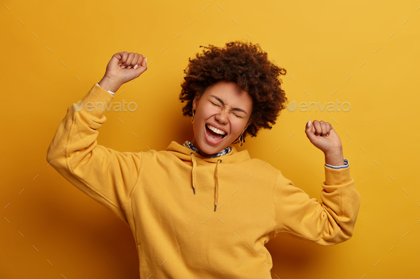 Yes I did it! Overjoyed optimistic woman with Afro hair, celebrates achievement, dances with raised