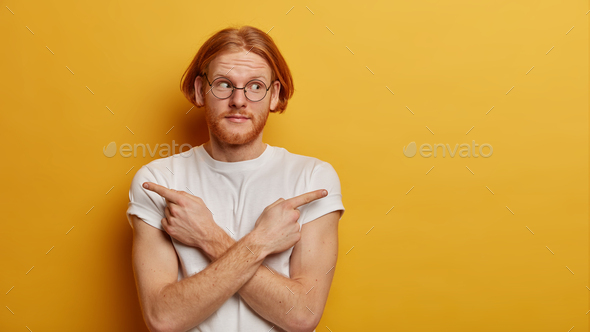 Confused youngster with red bob hair, beard, crosses arms over body and points sideways, chooses bet