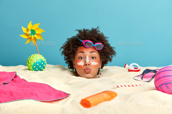 Outdoor shot of young curly woman in romantic mood, spends time at beach, applies sunscreen cream to