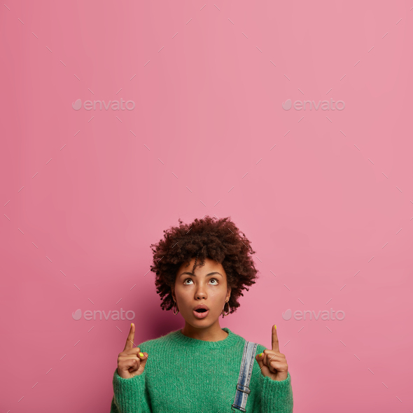 Confused shocked woman with Afro hairstyle, points upwards and says wow, promots product and discuss