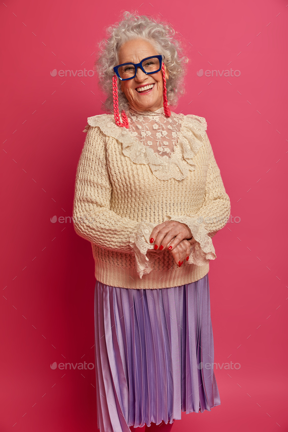 Positive wrinkled woman wears glasses, stylish clothes, smiles happily, expresses positive mood, bei