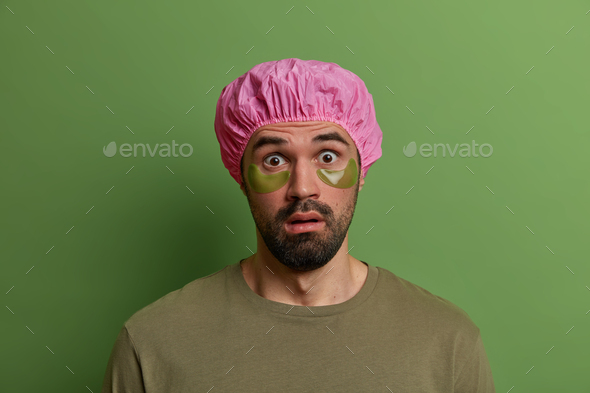 Young stunned unshaven European man stares with disbelief, finds out shocking news, wears bath cap a