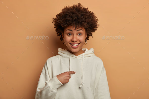 Surprised glad modest curly woman with gentle smile points at herself, asks about permission, wears