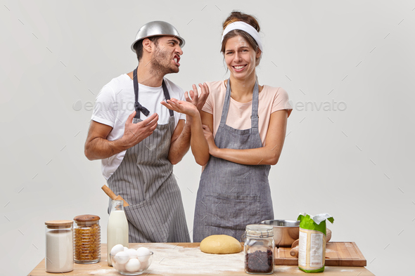 Annoyed adult man looks angrily at wife, asks to stop cooking, feels tired of making dough, cheerful