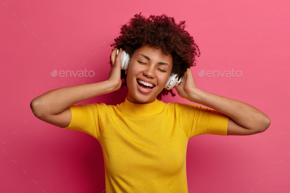 Carefree pleased millennial girl listens song in headphones, enjoys playlist, moves with rhythm, smi