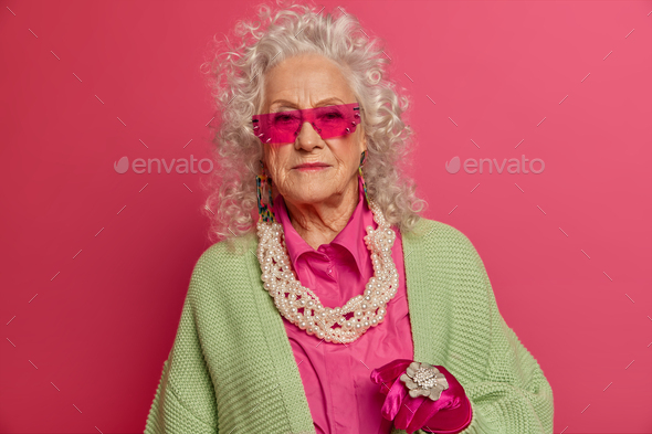 Senior woman wears trendy sunglasses, fashionable clothes and jewelry, has wrinkled face, curly grey