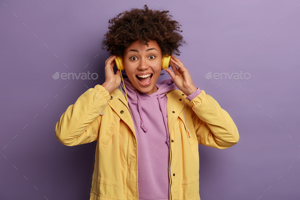 Photo of optimistic ethnic millennial girl listens songs in headphones, enjoys rhythm of music, laug