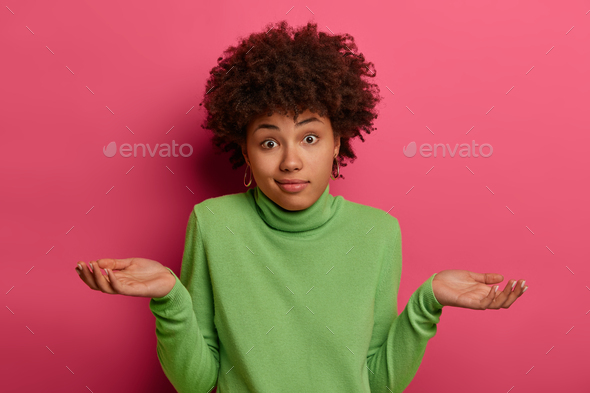 Indifferent confused black woman spreads palms confusingly, has doubts, wears green turtleneck, has