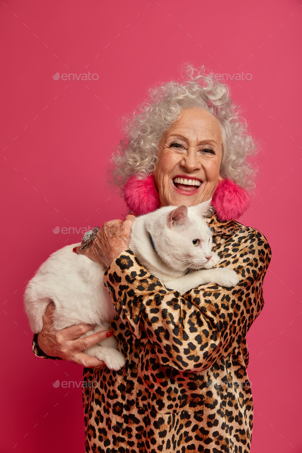 Pleased fashionable elderly woman holds domestic pet, feels not lonely in company of cat, dressed in