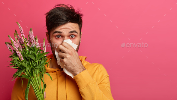 Unhealthy young man has blockage, blows nose, sufferd from allergy symptoms, covers nose with handke
