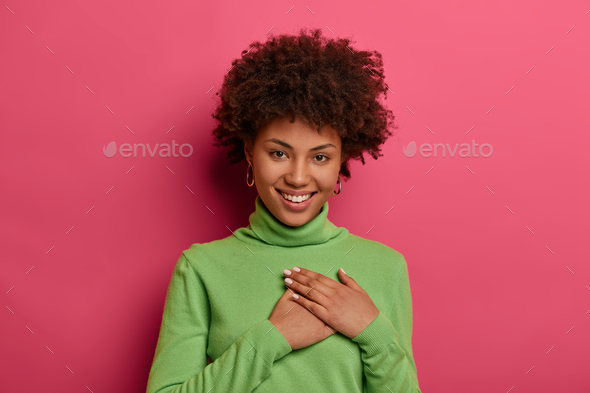 Positive young woman feels thankful, keeps hands pressed to heart, wears green turtleneck, smiles po