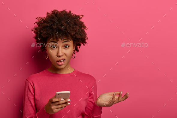 Distrubed dark skinned woman has problematic concerned face expression, holds smartphone, raises pal