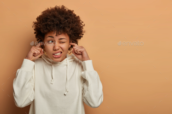 Stressed nervous curly woman plugs ears, annoyed with loud sound, asks stop making noise, clenches t