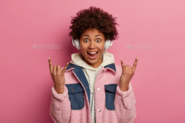 Excited dark skinned curly woman enjoys listening rock music in headphones, makes heavy metal gestur