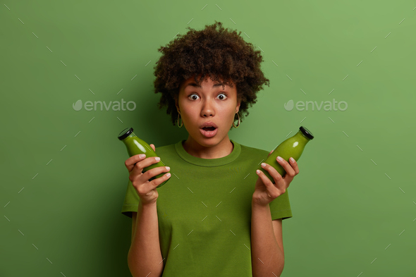 Attractive Afro American woman holds bottle of fresh blended green vegetable spinach smoothie, stare