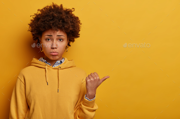 Portrait of dissatisfied dark skinned woman points away with thumb, gives negative feedback, reacts