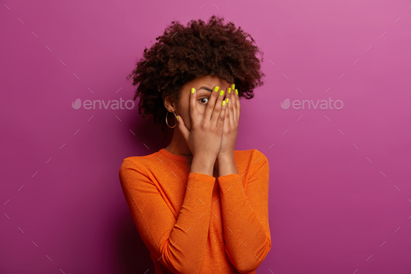 Young ethnic woman peeks through fingers, hides face with palms, has bright manicure, afraids of see