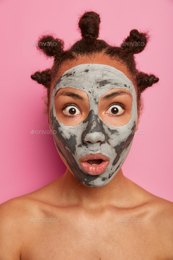Emotional shocked Afro American woman wears mask for face, reduces wrinkles on complexion, stands na
