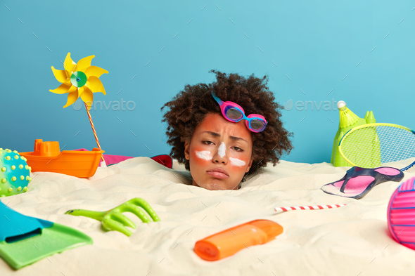 African American woman on vacation poses in sand, has upset and bored face expression, skin hurt fro