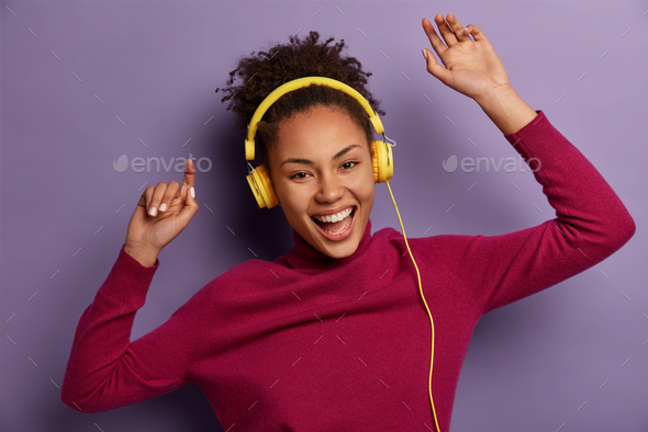 Portrait of excited curly haired woman dances carefree as listens audio track in headphones, enjoys