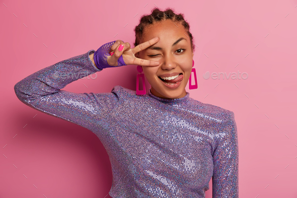 Half length shot of happy dark skinned woman makes peace disco gesture over eye, sticks out tongue,