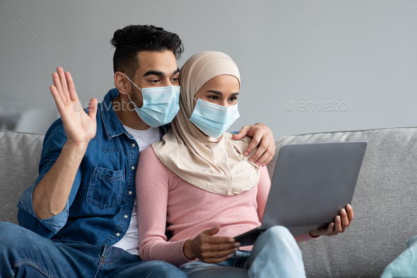 Muslim couple in face masks waving at laptop screen