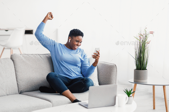 Excited black lady wearing headphones using phone