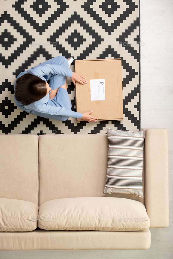 View of brunette girl in blue pajamas sitting by couch and going to open box