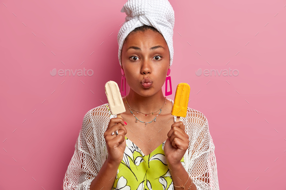 Lovely dark skinned model keeps lips folded, poses with vanilla and mango ice creams, eats unhealthy