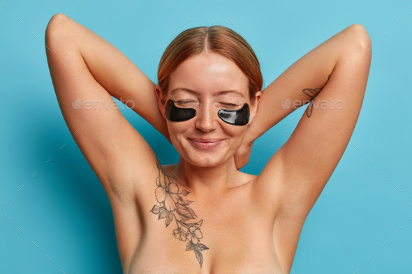 Beautiful sincere freckled woman keeps hands behind neck, closes eyes, applies black collagen patche