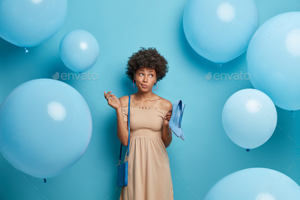 Thoughtful woman wears long beige dress, holds blue high heeled shoes to match bag, comes on friends