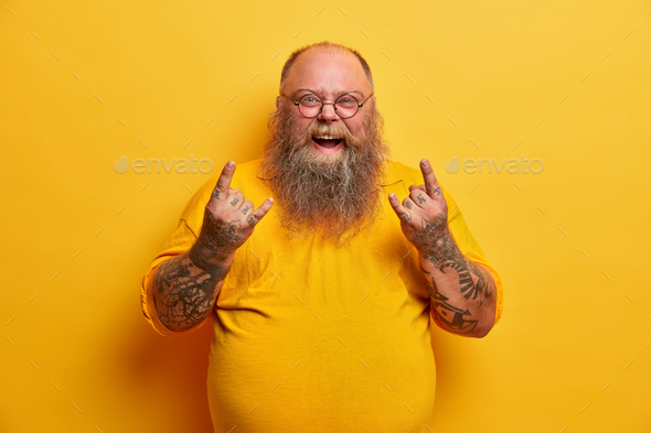 Obese funny man in yellow t shirt, shows heavy metal sign, attends concert of favorite music band, h