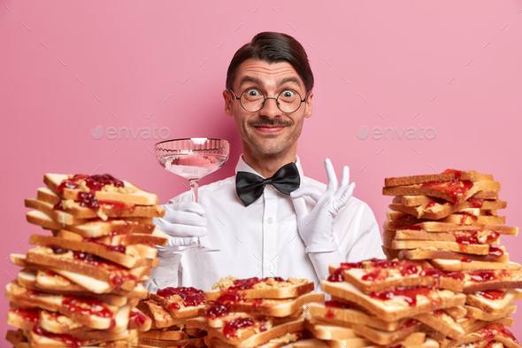Photo of cheerful waiter in uniform, poses with glass of martini, ready to take order from restauran