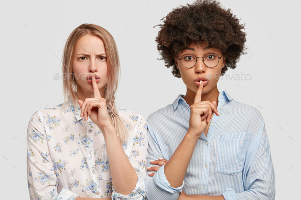 Two mixed race women demonstrate hush gesture, demand complete silence, ask to be mute and not make