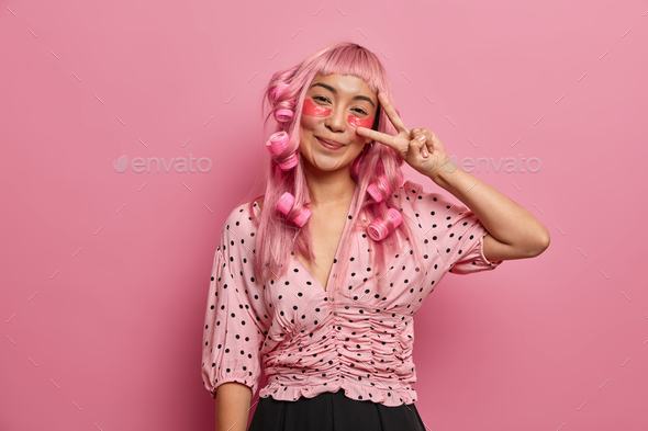 Happy pink haired girl applies eye patches for reducing dark circles, makes peace sign, wears hair c