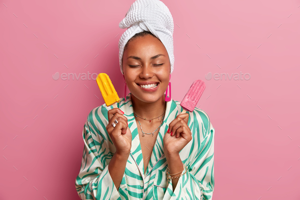 Pleased dark skinned woman bites lips, holds tasty appetizing ice cream, imagines pleasant taste of